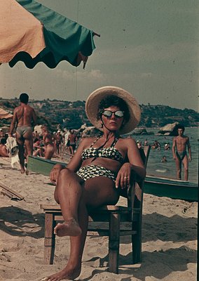 Seaside scene featuring a woman in a straw hat and patterned bikini posing on a wooden chair, 1960s beach culture. Crowds, bo...