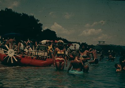 Vintage beach scene featuring a red inflatable raft with a sunburst logo, mid-20th century swimwear, and a lively crowd enjoy...