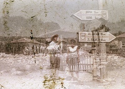 Vintage sepia-toned photo of rural roadside in Bulgaria, 1960s-70s. Two women in traditional attire pose near a rustic fence;...