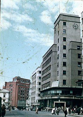 Mid-century urban street scene featuring a prominent **1960s-era apartment block** with a clock tower, flanked by smaller bri...