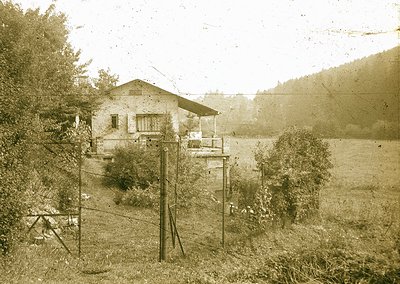 Vintage sepia-toned rural house with wooden balcony, surrounded by overgrown vegetation and utility poles. Likely early-to-mi...