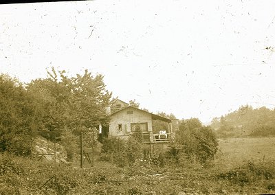 Vintage sepia-toned photo of a modest single-story house with a porch, surrounded by dense greenery and overgrown vegetation....