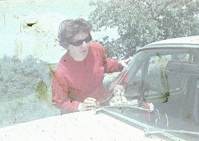 Vintage sepia-toned photo of a woman in a red sweater and sunglasses opening a classic car door, likely a 1960s–1970s model. ...