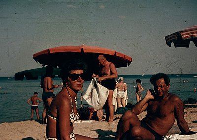 Vintage seaside scene with mid-20th-century beachgoers, likely 1960s-70s. Two men in swim trunks pose under a red umbrella; o...