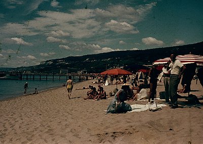 Vintage seaside scene with wooden pier extending into calm waters. Crowded sandy beach with mid-20th-century attire—swimsuits...