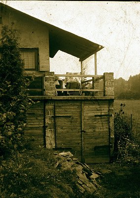 Vintage sepia-toned photo of a rustic wooden outbuilding with a small porch, likely a barn or storage shed. Open double doors...