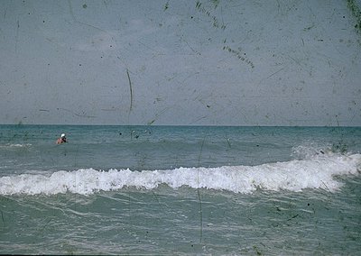 Vintage seaside shot featuring a lone swimmer in mid-wave. Faded, textured print suggests mid-20th century coastal photograph...