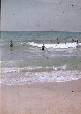 Vintage seaside scene with 1960s-style swimwear, groups wading in shallow waves. Beach appears sandy with visible footprints....