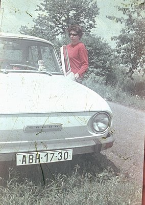 Vintage sepia-toned photo of a woman in a red sweater leaning on an open door of a **Škoda 100/110** (1960s–70s). License pla...