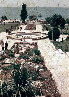 Vintage seaside promenade with symmetrical floral gardens, circular fountain, and wide pathways. Mid-20th century attire sugg...