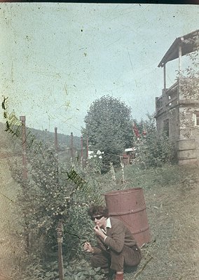 Vintage sepia-toned photo of a person kneeling in a lush garden, using an early 20th-century telephone. Wooden fence, rustic ...