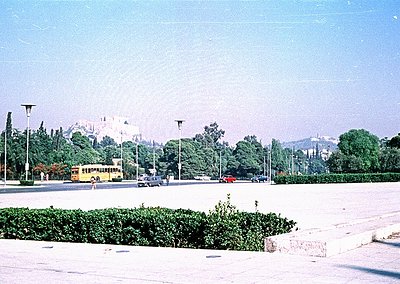 Vintage urban scene featuring a yellow bus and classic cars on a wide, tree-lined boulevard. Prominent historic castle ruins ...