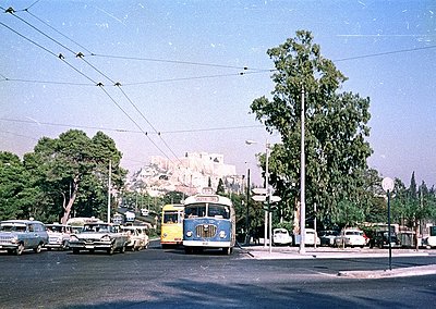 Vintage 1950s-60s urban scene featuring a blue & yellow trolleybus at a busy intersection. Classic cars, overhead wires, and ...