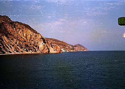 Vintage coastal scene featuring rugged, layered cliffs meeting deep blue waters. Partial view of a green lifebuoy on right su...