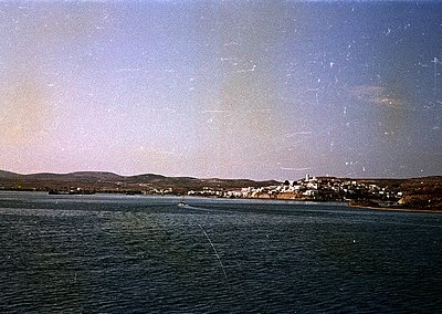 Vintage coastal town framed by calm waters, likely Mediterranean. Low-lying buildings with flat roofs and whitewashed walls l...