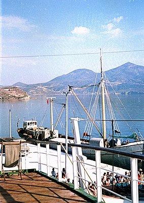Vintage ferry docked at coastal port, with passengers on deck overlooking deep blue waters and mountainous coastline. Mid-20t...