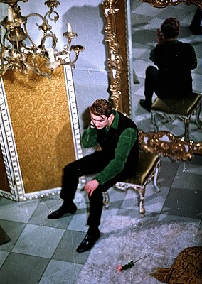 Young man in 1960s-70s streetwear (green sweater, dark pants, boots) sits on ornate marble floor in a grand, mirrored room. G...
