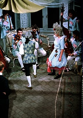 Vintage indoor ballroom scene featuring a man in 19th-century formalwear (tuxedo with floral vest) dancing with a woman in a ...