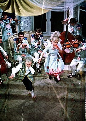 Vintage 1970s indoor folk dance with musicians playing traditional instruments—violins, accordion, cello. Couple in mid-dance...