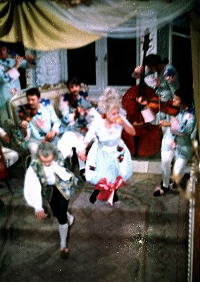 Vintage indoor gathering featuring children in 1960s-70s folk costumes dancing to live string music. White tablecloths, flora...