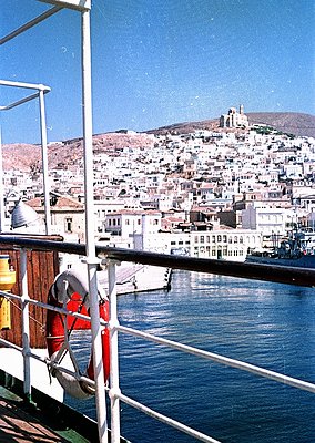 Coastal cityscape viewed from a ferry, featuring densely packed Mediterranean-style buildings with flat roofs and whitewashed...