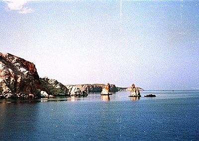 Vintage coastal scene featuring jagged volcanic rock formations rising from calm, reflective waters. Low-angle sunlight casts...