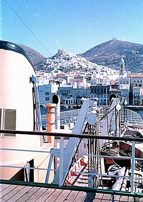 Mid-20th century ferry deck overlooking coastal city with hilltop fortress. White railings, vintage lifebuoys, and rope barri...