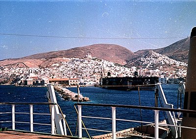 Vintage coastal town nestled on rocky terrain, viewed from a ferry. Dense cluster of whitewashed buildings cascades down hill...