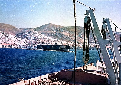 Vintage port scene featuring a mid-20th century cargo ship docked near coastal cityscape. Industrial cranes and ropes dominat...