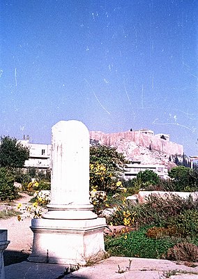 Ancient Greek-style column fragment in a sunlit archaeological park, set against the Acropolis hill in Athens. Weathered marb...