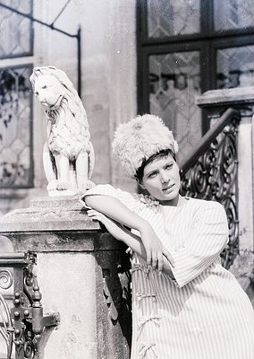 Vintage black-and-white portrait of a woman in early 20th-century fashion, posing beside a marble lion statue atop ornate sto...