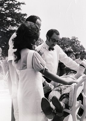 Black-and-white photo of a 1960s outdoor gathering: a woman in a sleeveless blouse adjusts a seated man’s tie while others ob...
