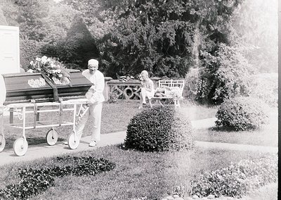 Mid-20th century funeral procession in a landscaped cemetery. A man in formal attire pushes a hearse with floral arrangements...