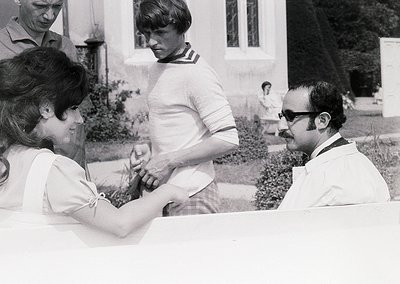 Black-and-white photo capturing a 1970s medical exam scene. A female patient in a short-sleeved blouse has her arm extended f...