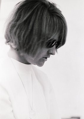 Portrait of a woman with a short, voluminous 1960s hairstyle, wearing round sunglasses and a white blouse with a pendant neck...