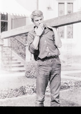 Young man in 1960s-style casual attire—rolled-up sleeves, button-down shirt, and loose-fitting trousers—standing outdoors nea...