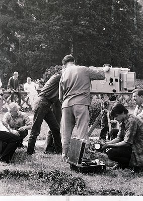 Mid-20th century film crew operating vintage 16mm camera setup outdoors. Crew members in casual 1950s-60s attire—plaid shirts...