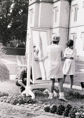 Vintage black-and-white photo of three women in 1960s-era attire posing with a large handheld mirror outdoors. The woman knee...