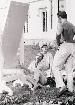 Vintage black-and-white photo of three individuals posing with a large, open, white folding screen in an outdoor setting. The...