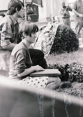 Black-and-white street scene from the 1960s–70s, featuring a young man seated on a low wall, engrossed in a book. He wears a ...