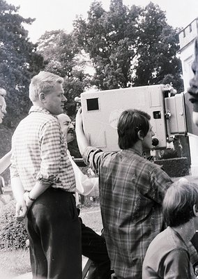 Vintage film crew operating a large, boxy 16mm/Super 8 handheld camera (, , ). Three men in plaid shirts and casual attire di...