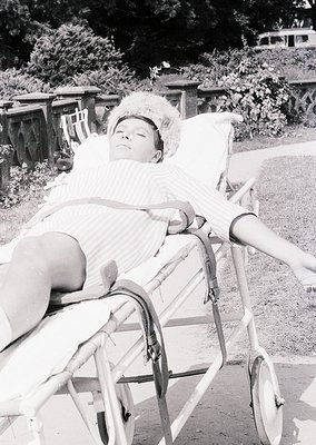Mid-20th century baby reclining in vintage metal pram on paved path, surrounded by lush greenery and wooden fencing. Classic ...
