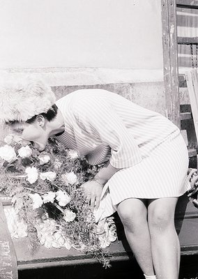 A woman in a 1960s-style striped dress arranges a floral wreath, likely for a memorial or funeral. The setting appears to be ...