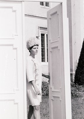 Mid-century woman in a fitted, floral-patterned dress and voluminous bouffant hairstyle poses beside a white door with vertic...