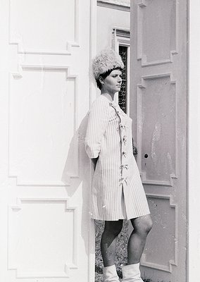 Vintage black-and-white portrait of a woman in a striped housecoat and fur hat, leaning against a paneled door. The architect...