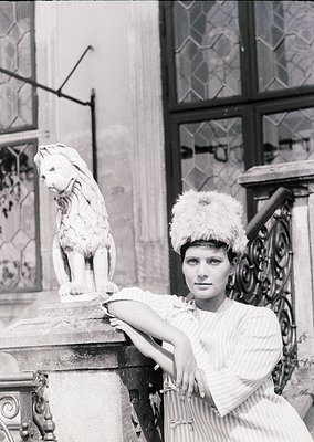 Black-and-white portrait of a woman in 1950s-60s fashion, posing beside a marble lion statue in front of ornate wrought-iron ...