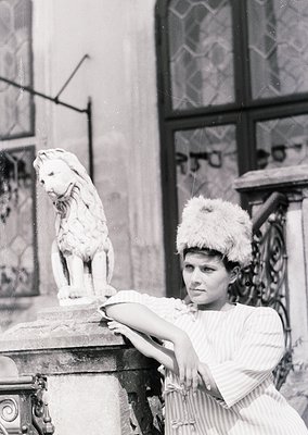 Vintage black-and-white portrait of a woman in 1920s-30s fashion, posing beside a marble lion statue atop an ornate pedestal....