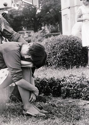 Black-and-white candid of a seated man in 1960s-era clothing (plaid shirt, rolled trousers) adjusting his tie outdoors. Lush ...