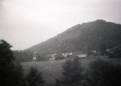 Vintage black-and-white rural landscape featuring modest wooden houses nestled at the base of a forested hillside. Fog or mis...