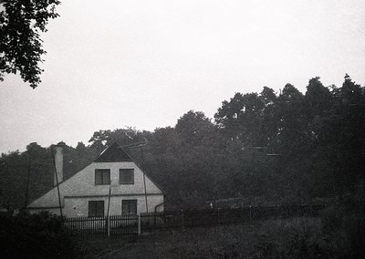 Vintage black-and-white photo of a modest, two-story farmhouse with a gabled roof and chimney, surrounded by dense forest. Fe...